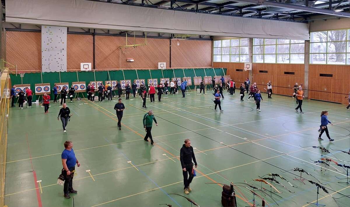 Schulsporthalle, im Hintergrund sind 16 Zielscheiben eng nebeneinander zu sehen, auf jede Scheibe schie&szlig;en 2 Sch&uuml;tzen, Bogensch&uuml;tzen stehen vor den Scheiben und notieren ihre Ringe, einige sind auf dem Weg zur Startlinie, im Vordergrund stehen B&ouml;gen aller Art auf dem Boden.