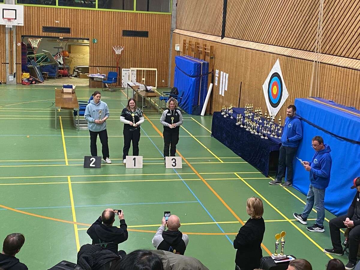 Schulsporthalle, im Hintergrund steht ein Garagentor offen, davor stehen 2 Tische, rechts sieht man Pokale auf einem Tisch mit einer Stufe der mit samtenem blauen Stoff bedeckt ist, dar&uuml;ber h&auml;ngt an der Wand eine gro&szlig;e Zielscheibe der Bodensch&uuml;tzen, in der Mitte des Bildes stehen auf dem Boden die Ziffen 2, 1, 3 als Schilder und dahinter stehen drei Frauen mit Pokalen in der Hand.